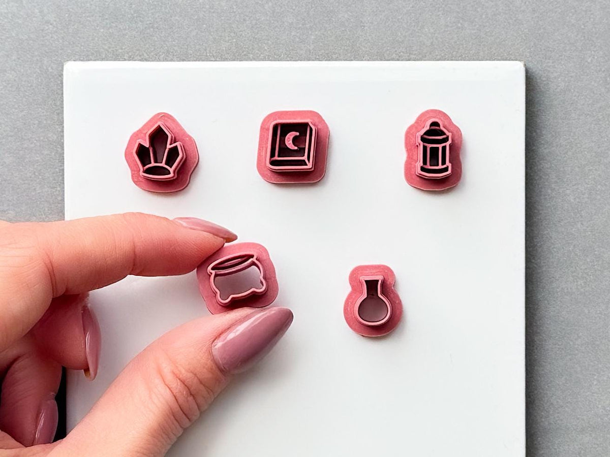 Five miniature witchy clay cutters displayed on a white surface, featuring a witch’s hat, spell book with crescent moon, lantern, cauldron and potion bottle. The cauldron cutter is held between fingers to show its size and detailed design. Gothic, witchy and Halloween-inspired clay cutter set.