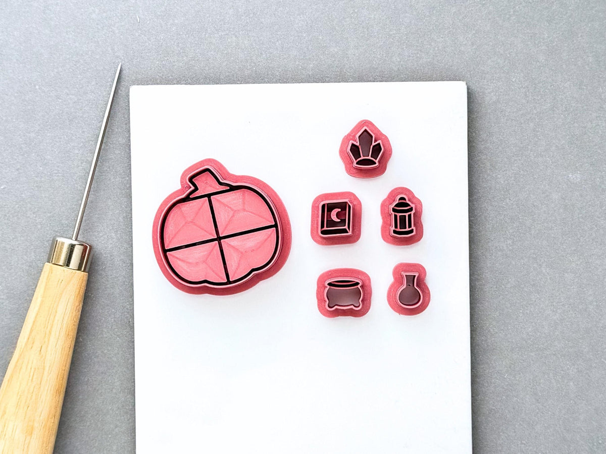 Halloween pumpkin-shaped bookcase clay cutter with miniature witchy cutters, including a witch’s hat, spell book, lantern, cauldron and potion bottle. Pink 3D-printed cutters displayed together on a white surface with a clay sculpting tool. Gothic, witchy and Halloween-inspired design.