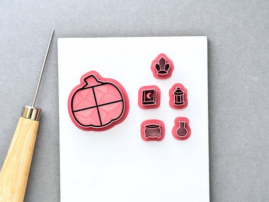 Halloween pumpkin-shaped bookcase clay cutter with miniature witchy cutters, including a witch’s hat, spell book, lantern, cauldron and potion bottle. Pink 3D-printed cutters displayed together on a white surface with a clay sculpting tool. Gothic, witchy and Halloween-inspired design.