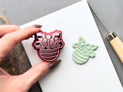 Christmas bauble polymer clay cutter held between fingers beside a matching finished clay cut-out, showcasing the detailed snowflake bow, floral centre and striped bauble design.