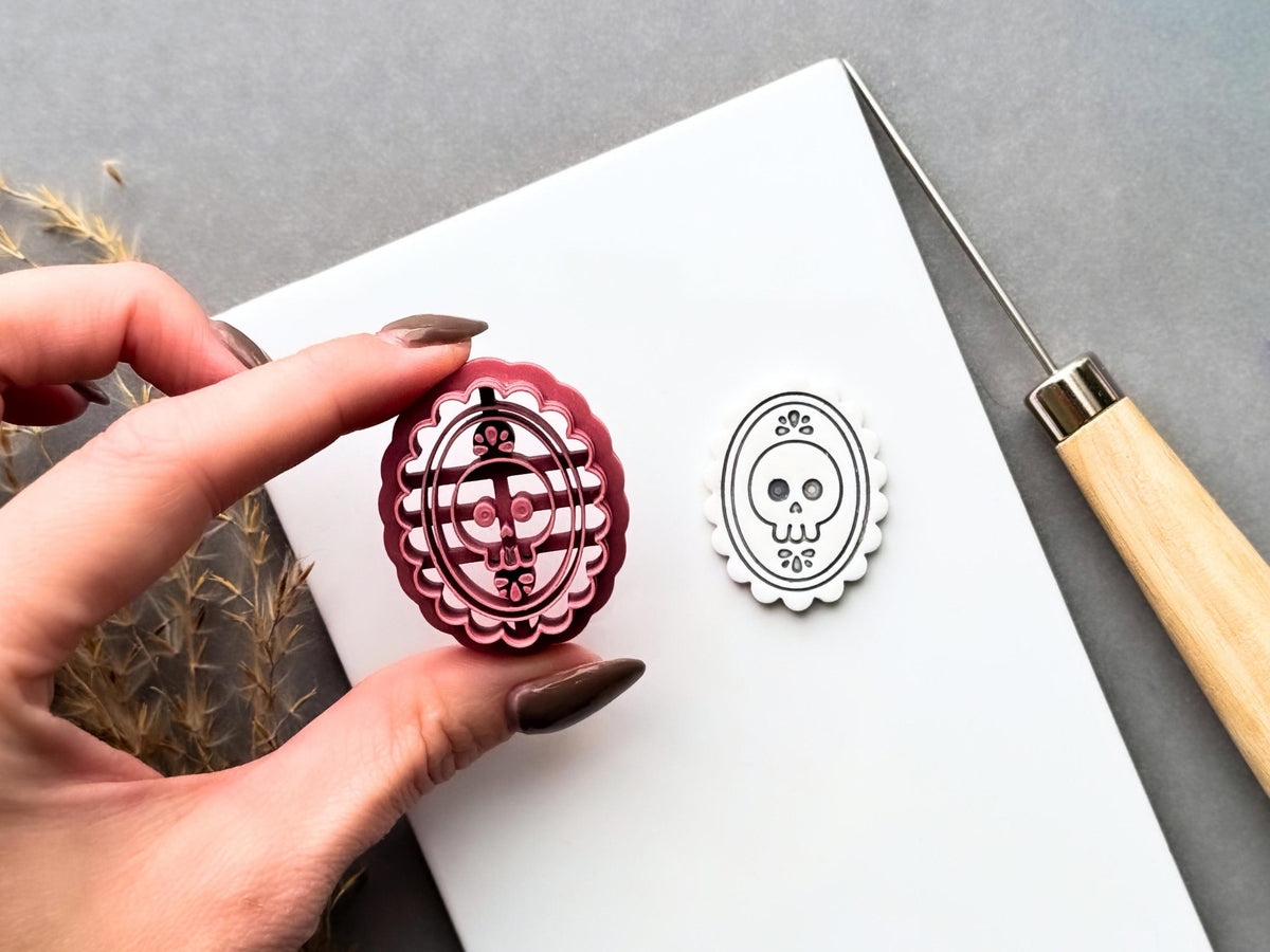 Gothic skull cameo polymer clay cutter held between fingers beside a matching finished clay cut-out, showcasing the detailed skull with floral accents, ornate oval frame and scalloped edge.