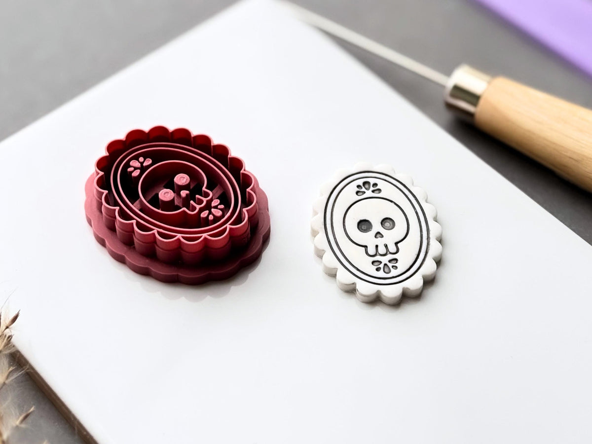 Gothic skull cameo polymer clay cutter displayed beside a matching finished clay cut-out, showcasing the raised skull and floral stamp details within an ornate oval frame and scalloped border. Angled view highlighting the deep cutting walls and internal stamp structure.