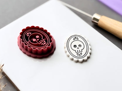 Gothic skull cameo polymer clay cutter displayed beside a matching finished clay cut-out, showcasing the raised skull and floral stamp details within an ornate oval frame and scalloped border. Angled view highlighting the deep cutting walls and internal stamp structure.