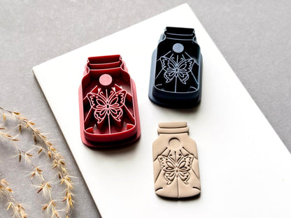 Butterfly specimen jar clay cutters in red and blue, featuring detailed symmetrical butterfly designs with decorative floral details, shown alongside a finished cream polymer clay butterfly cut-out. Different cutter colours and sizes are displayed on a white surface.