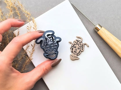 Medusa head polymer clay cutter held between fingers for scale, showcasing the detailed facial features, intricate snake-shaped hair and fine cutting edge, beside a matching finished cream polymer clay Medusa embellishment.