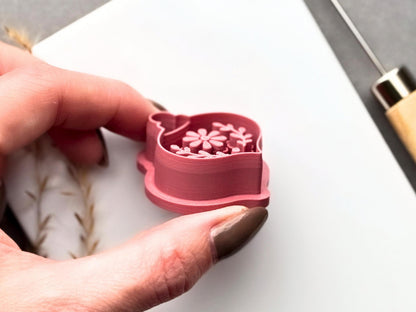 Side view of a floral Christmas bauble clay cutter held in a hand, showing the raised cutting edge, layered 3D construction and strong support wall. The central flower and leafy botanical stamp details are visible inside the cutter.