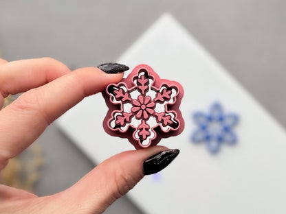Snowflake polymer clay cutter held between fingers, showcasing the detailed six-point snowflake design with floral centre and branch-like decorative details.
