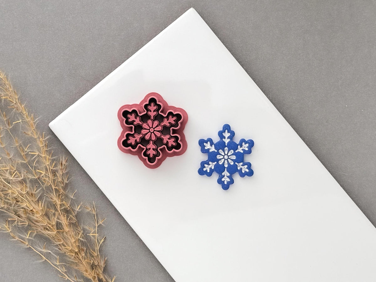 Snowflake polymer clay cutter displayed beside a matching finished clay cut-out, featuring a detailed six-point snowflake with floral and branch-like decorative details.