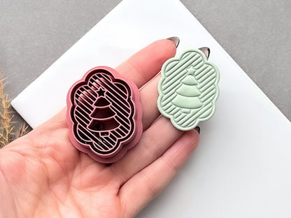 Pink polymer clay cutter shaped like a scalloped Christmas tree with diagonal striped detail and a small star at the top, shown beside a pale green finished clay tree held in a hand.