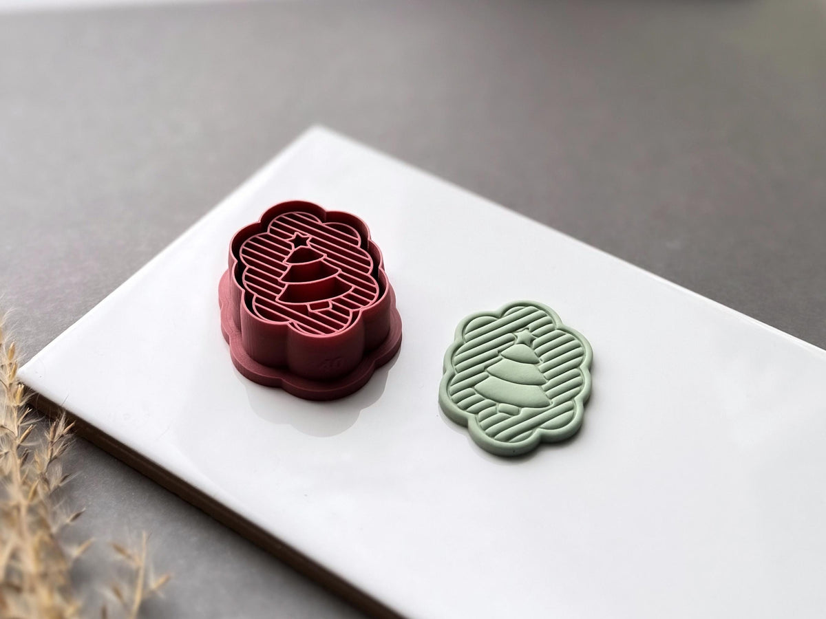 Christmas tree polymer clay cutter displayed beside a matching finished clay cut-out, featuring a scalloped oval frame, striped background and layered Christmas tree design. Angled view showcasing the raised stamp details and deep cutting walls.