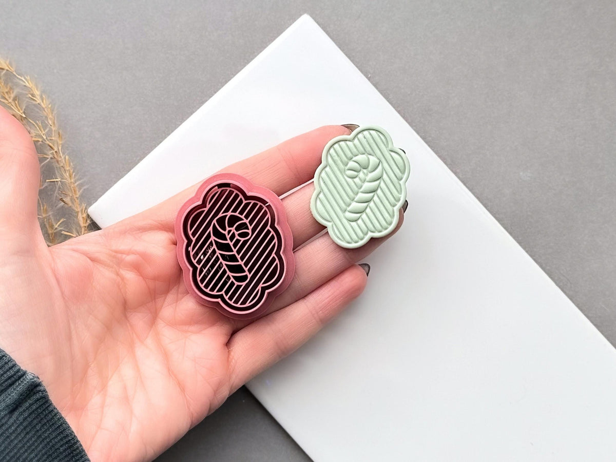 Christmas candy cane polymer clay cutter held in a hand beside a matching finished clay cut-out, featuring a scalloped frame, striped background and central candy cane design.