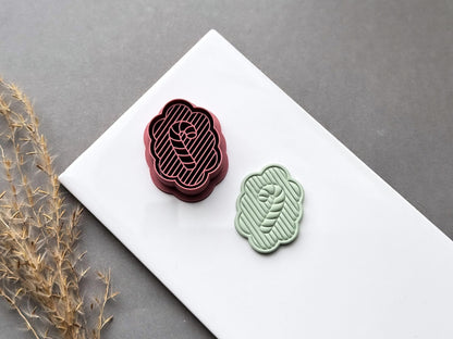 Dark pink polymer clay cutter with scalloped frame, horizontal stripes and candy cane inner stamp lines next to a sage coloured clay cut out earring.
