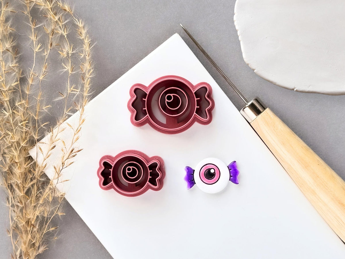 Eyeball-shaped candy clay cutters displayed in two sizes, featuring a central eyeball design within a wrapped candy shape, shown beside a finished white polymer clay eyeball candy with purple twisted ends. Halloween-inspired design displayed on a white surface.