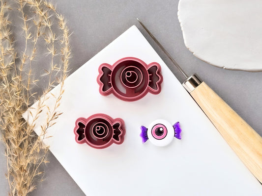 Eyeball-shaped candy clay cutters displayed in two sizes, featuring a central eyeball design within a wrapped candy shape, shown beside a finished white polymer clay eyeball candy with purple twisted ends. Halloween-inspired design displayed on a white surface.