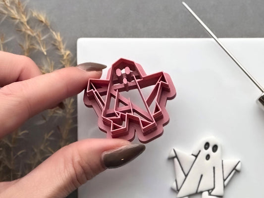 Origami ghost polymer clay cutter held in a hand, showcasing the folded geometric design, three facial details and raised cutting edge, with a finished white origami ghost cut-out in the background.
