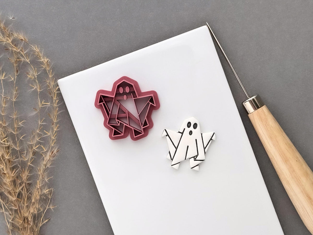 Origami paper ghost shaped polymer clay cutter featuring a folded geometric ghost design with facial details, shown beside a finished white polymer clay origami ghost cut-out with black line details. Pink 3D-printed cutter displayed on a white surface.
