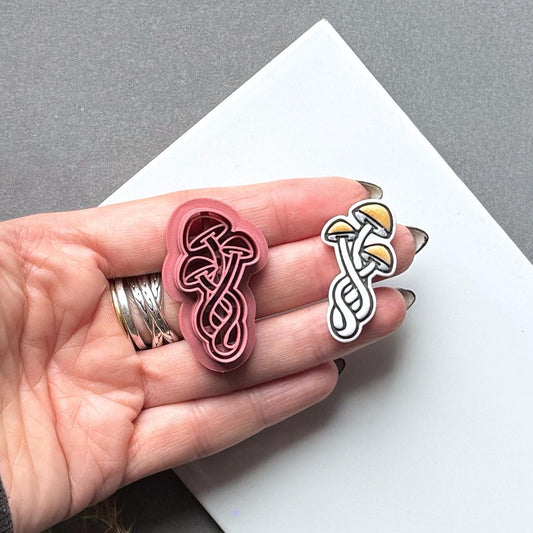 Triple mushroom polymer clay cutter and matching finished clay cut-out held in a hand for scale, featuring three overlapping mushrooms with curved stems and detailed cap outlines.