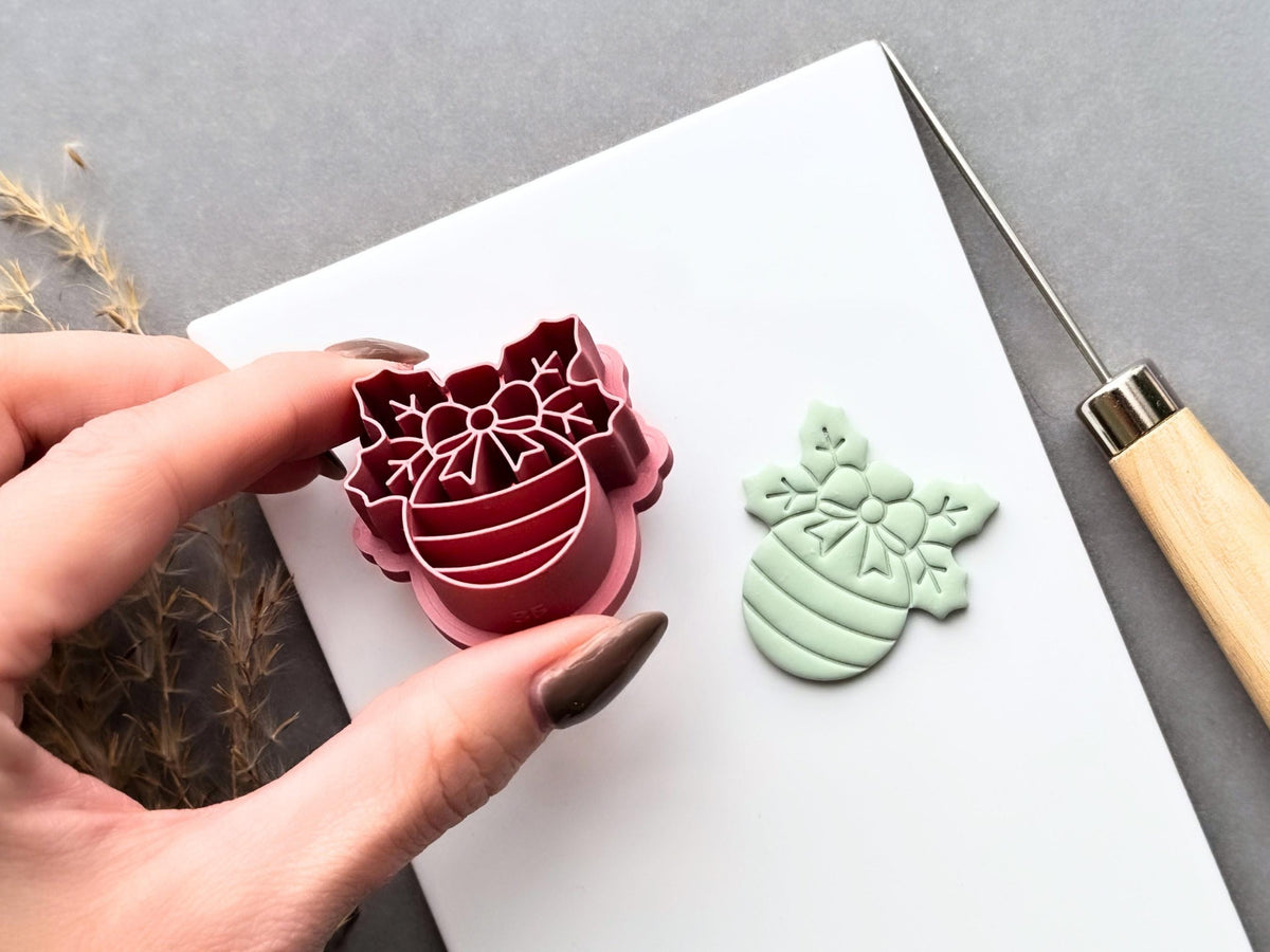 Christmas bauble polymer clay cutter held at an angle to showcase the raised snowflake bow and striped bauble stamp details, deep cutting walls and sturdy support structure, with the matching finished clay cut-out beside it.