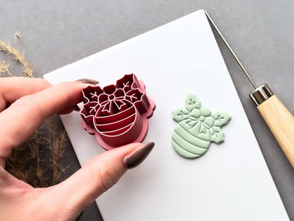 Christmas bauble polymer clay cutter held at an angle to showcase the raised snowflake bow and striped bauble stamp details, deep cutting walls and sturdy support structure, with the matching finished clay cut-out beside it.