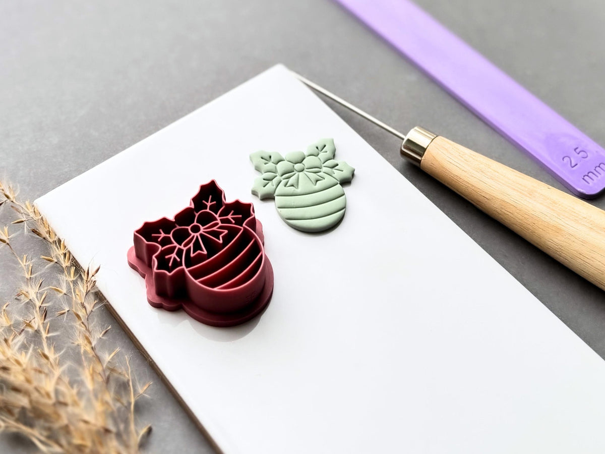 Christmas bauble polymer clay cutter displayed beside a matching finished clay cut-out, featuring a decorative snowflake bow, floral centre and striped bauble design. Angled view showcasing the raised stamp details, deep cutting walls and sturdy support structure.