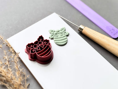 Christmas bauble polymer clay cutter displayed beside a matching finished clay cut-out, featuring a decorative snowflake bow, floral centre and striped bauble design. Angled view showcasing the raised stamp details, deep cutting walls and sturdy support structure.