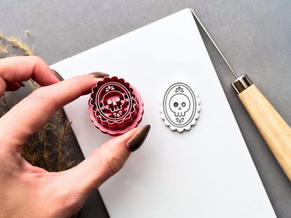 Gothic skull cameo polymer clay cutter held at an angle to showcase the raised skull and floral stamp details, ornate oval frame, scalloped cutting edge and internal structure, with the matching finished clay cut-out beside it.