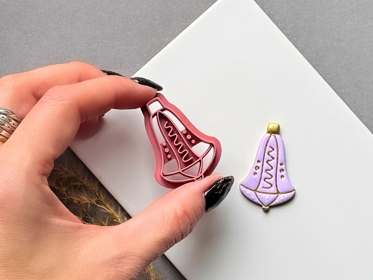 Christmas bell polymer clay cutter held between fingers to showcase the detailed hanging bell shape, wavy central stamp motif and dotted accents, with a matching finished lavender clay embellishment featuring gold details beside it.