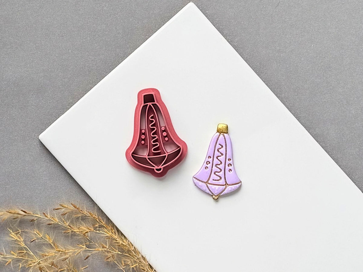 Christmas bell polymer clay cutter featuring a decorative hanging bell with a wavy central motif and dotted details, displayed beside a matching finished clay embellishment in lavender with gold accents.