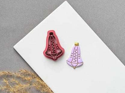 Christmas bell polymer clay cutter featuring a decorative hanging bell with a wavy central motif and dotted details, displayed beside a matching finished clay embellishment in lavender with gold accents.