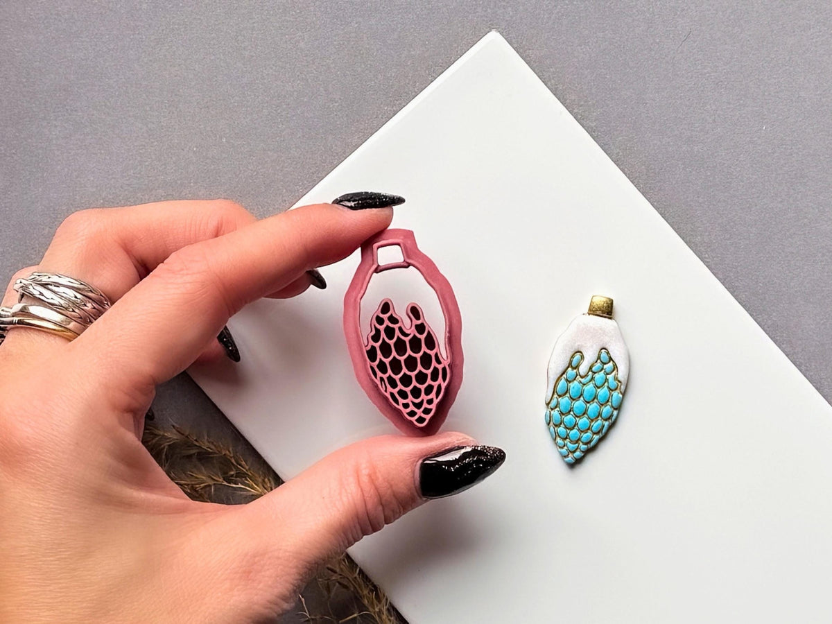 Christmas bauble polymer clay cutter held between fingers to showcase the elongated ornament shape and detailed scalloped scale stamp pattern, with a matching finished turquoise and gold clay embellishment beside it.