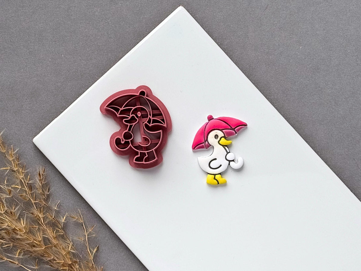 Umbrella polymer clay cutter featuring a cute duck character holding a curved umbrella, displayed beside a matching finished clay embellishment on a white tile.