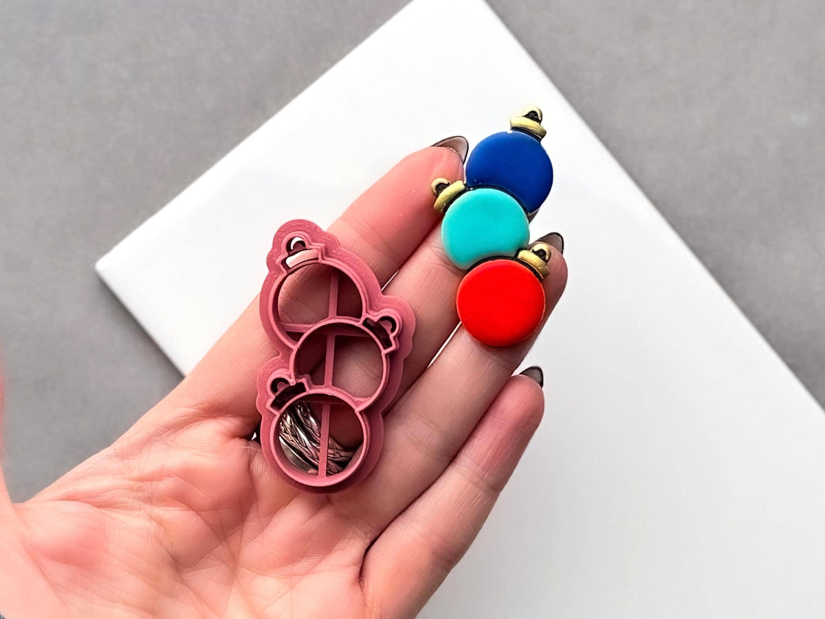 Christmas bauble trio polymer clay cutter held in a hand beside a finished clay embellishment featuring three stacked round baubles with decorative caps, showcasing the cutter’s raised cutting edge and internal support structure.
