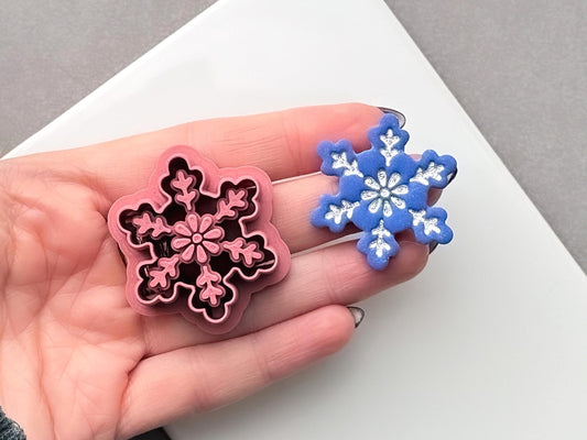Snowflake polymer clay cutter held in a hand beside a matching finished clay cut-out, featuring a detailed six-point snowflake with floral and branch-like decorative details.