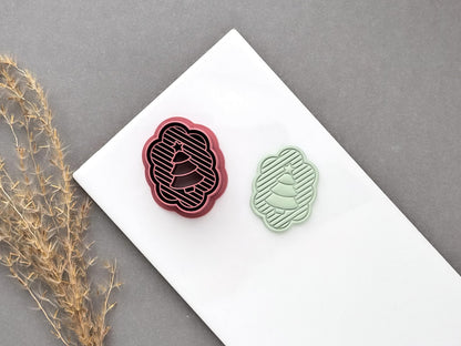 Christmas tree polymer clay cutter displayed beside a matching finished clay cut-out, featuring a scalloped oval frame, striped background and layered Christmas tree design.