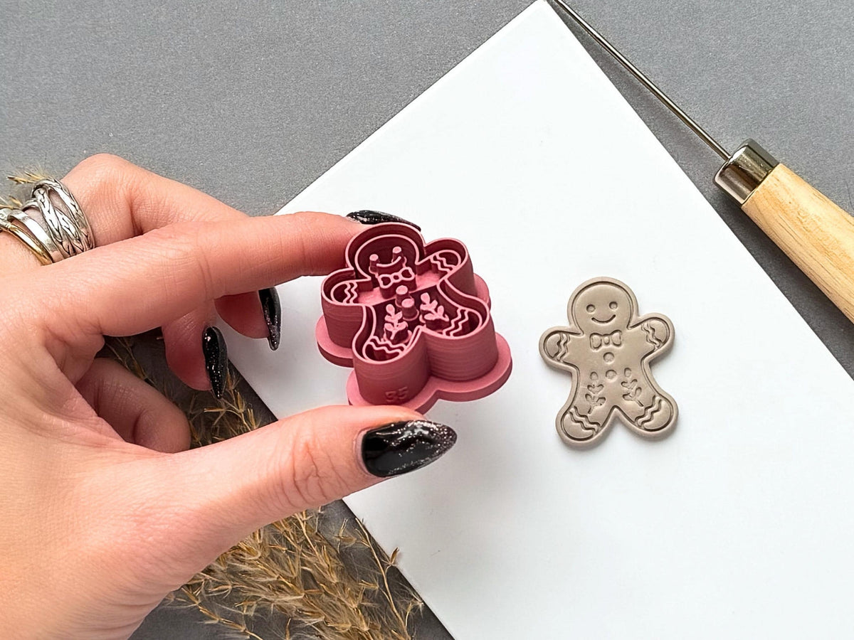 Gingerbread man polymer clay cutter held between fingers to showcase the raised cutting walls and detailed stamp design, with a matching finished gingerbread man clay embellishment displayed on a white tile.