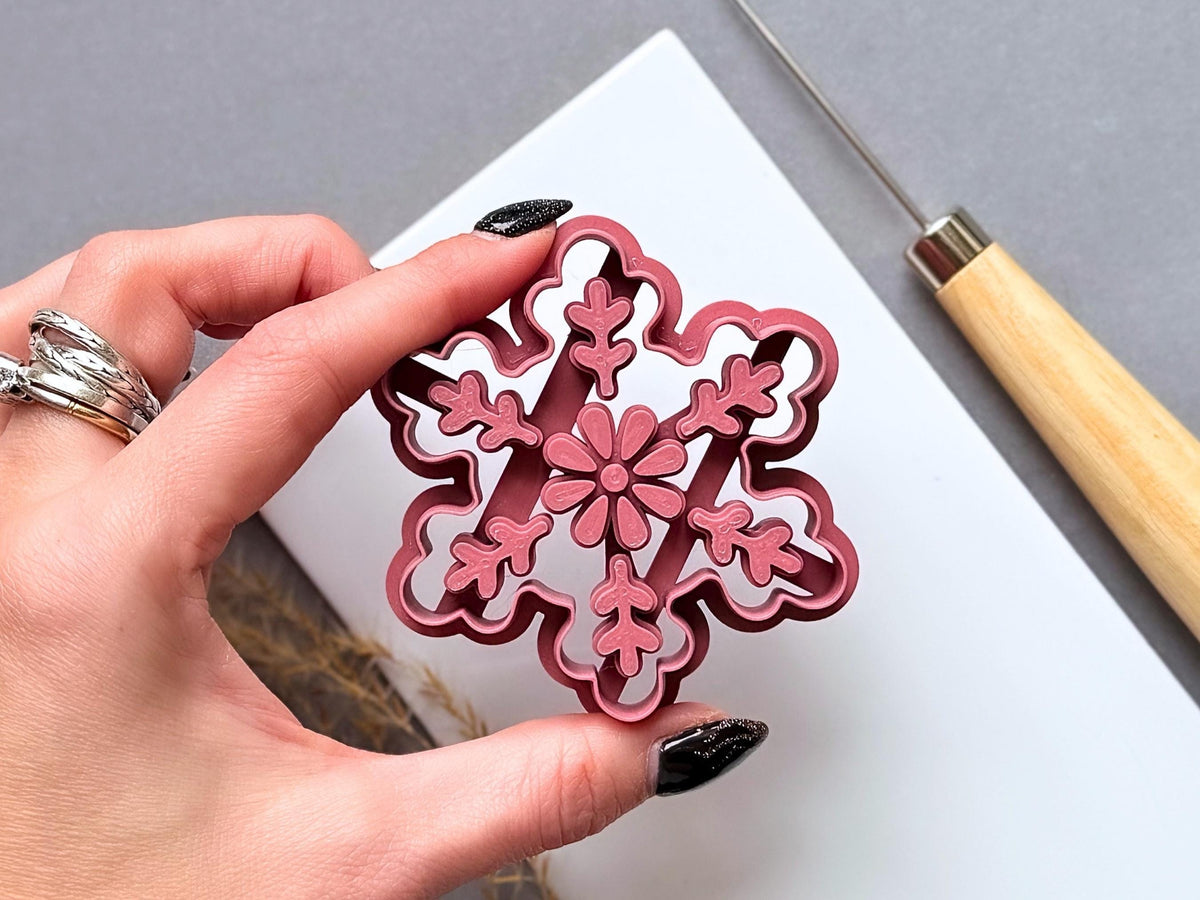 Large snowflake polymer clay ornament cutter held in a hand for scale, featuring a central flower and six decorative branches with leaf-like details, showcasing the intricate stamp design and shaped cutting edge.