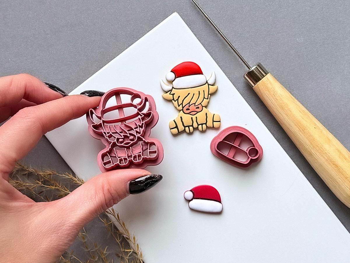 Highland cow Christmas polymer clay cutter and separate Santa hat cutter displayed beside a finished clay Highland cow wearing a Santa hat, with both cutters shown in close-up.