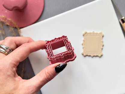 Vintage-style rectangular frame polymer clay cutter held in a hand for scale, featuring an ornate scalloped border with decorative loop and bead details, alongside a matching finished clay frame.