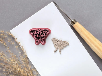 Moth polymer clay cutter displayed beside a matching finished clay moth, featuring delicate symmetrical wings, decorative botanical details and a long, flowing tail design.