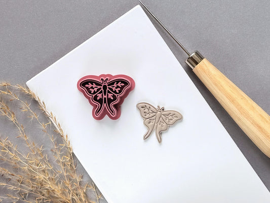 Moth polymer clay cutter displayed beside a matching finished clay moth, featuring delicate symmetrical wings, decorative botanical details and a long, flowing tail design.