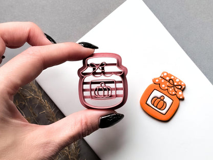 Halloween pumpkin jar polymer clay cutter held between fingers to showcase the detailed striped jar design, central pumpkin stamp and decorative bow lid, displayed beside the matching finished clay cut-out.