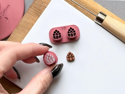 Pine cone polymer clay stamp cutter set featuring detailed scale patterns, displayed in a two-piece design with a matching finished clay pine cone embellishment and a hand-held cutter for close-up detail.