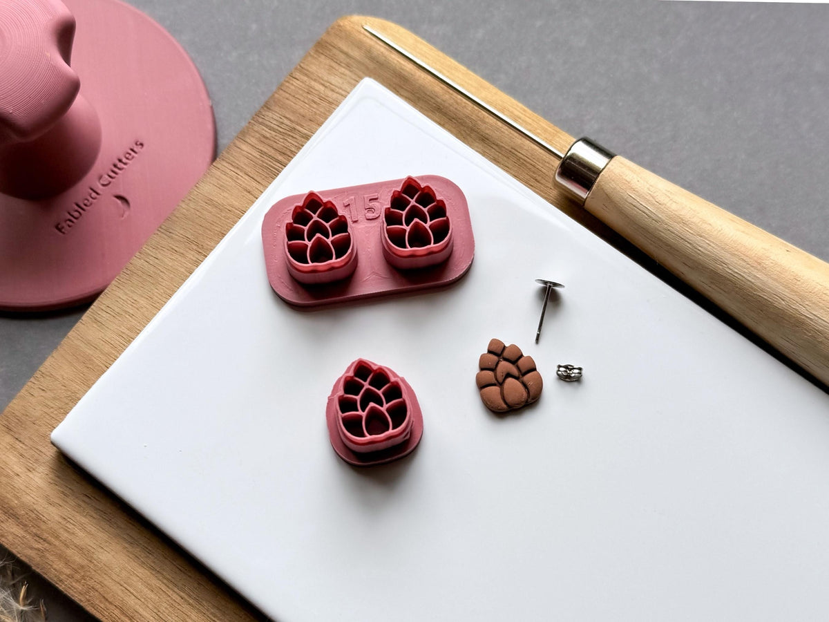 Pine cone polymer clay stamp cutter set featuring two detailed pine cone designs on a joined holder, alongside a separate single cutter and a matching finished clay pine cone embellishment. Small metal findings are displayed nearby for scale.