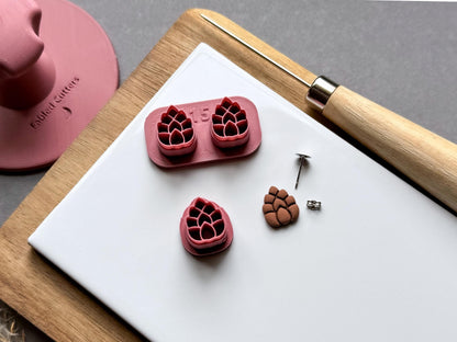 Pine cone polymer clay stamp cutter set featuring two detailed pine cone designs on a joined holder, alongside a separate single cutter and a matching finished clay pine cone embellishment. Small metal findings are displayed nearby for scale.