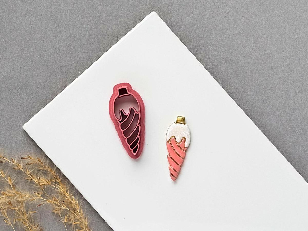 Christmas bauble polymer clay cutter featuring an elongated teardrop ornament with a flowing spiral stamp design, displayed beside a matching finished clay embellishment in coral pink.
