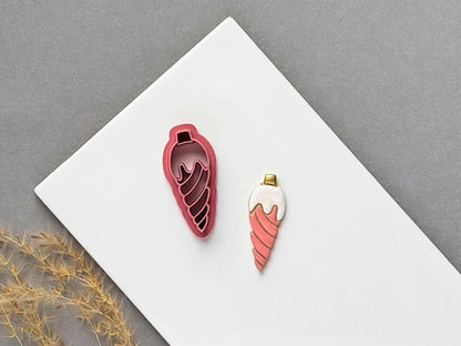 Christmas bauble polymer clay cutter featuring an elongated teardrop ornament with a flowing spiral stamp design, displayed beside a matching finished clay embellishment in coral pink.