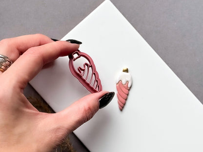 Christmas bauble polymer clay cutter held between fingers to showcase the elongated teardrop shape and flowing spiral stamp design, with a matching finished clay embellishment in coral pink and white in the background.
