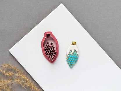 Christmas bauble polymer clay cutter featuring an elongated ornament with a decorative scalloped scale pattern, displayed beside a matching finished clay embellishment in turquoise and gold.
