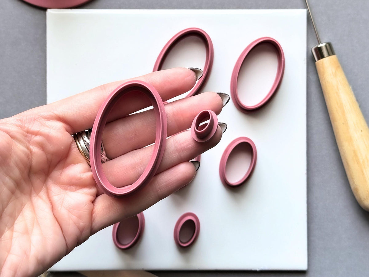 Pink oval polymer clay cutters displayed in multiple sizes, with one large oval cutter held in a hand and smaller cutters arranged on a white tile.