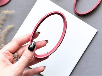 Large oval polymer clay cutter held between fingers to showcase its elongated shape, smooth inner outline and raised cutting wall, with another oval cutter visible in the background.